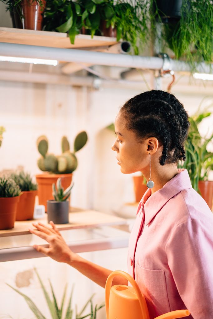 Woman in pink shirt observes potted plants on shelves indoors, holding an orange watering can.